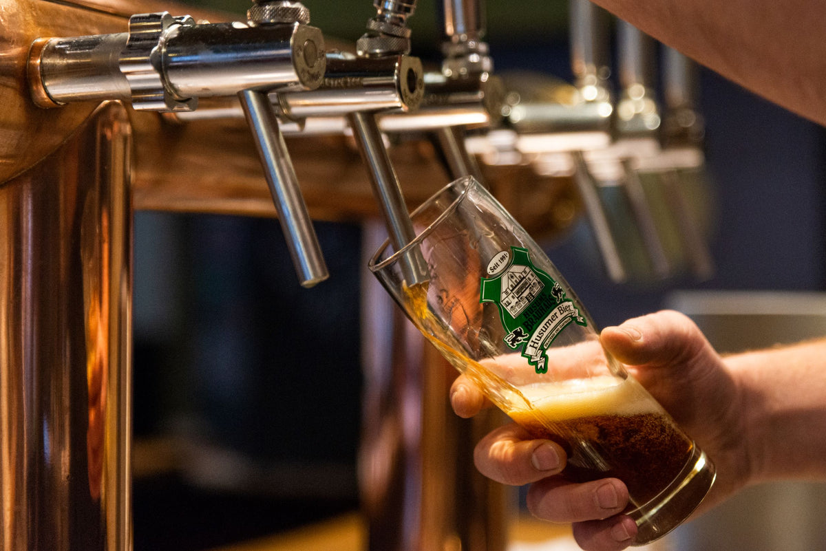 Das Foto zeigt den Zapfhahn aus dem Brauhaus. Man sieht das Glas in welches jemand das Bier zapft. Man erkennt nur die Hände die das Glas halten und den Zapfhahn bedienen. 