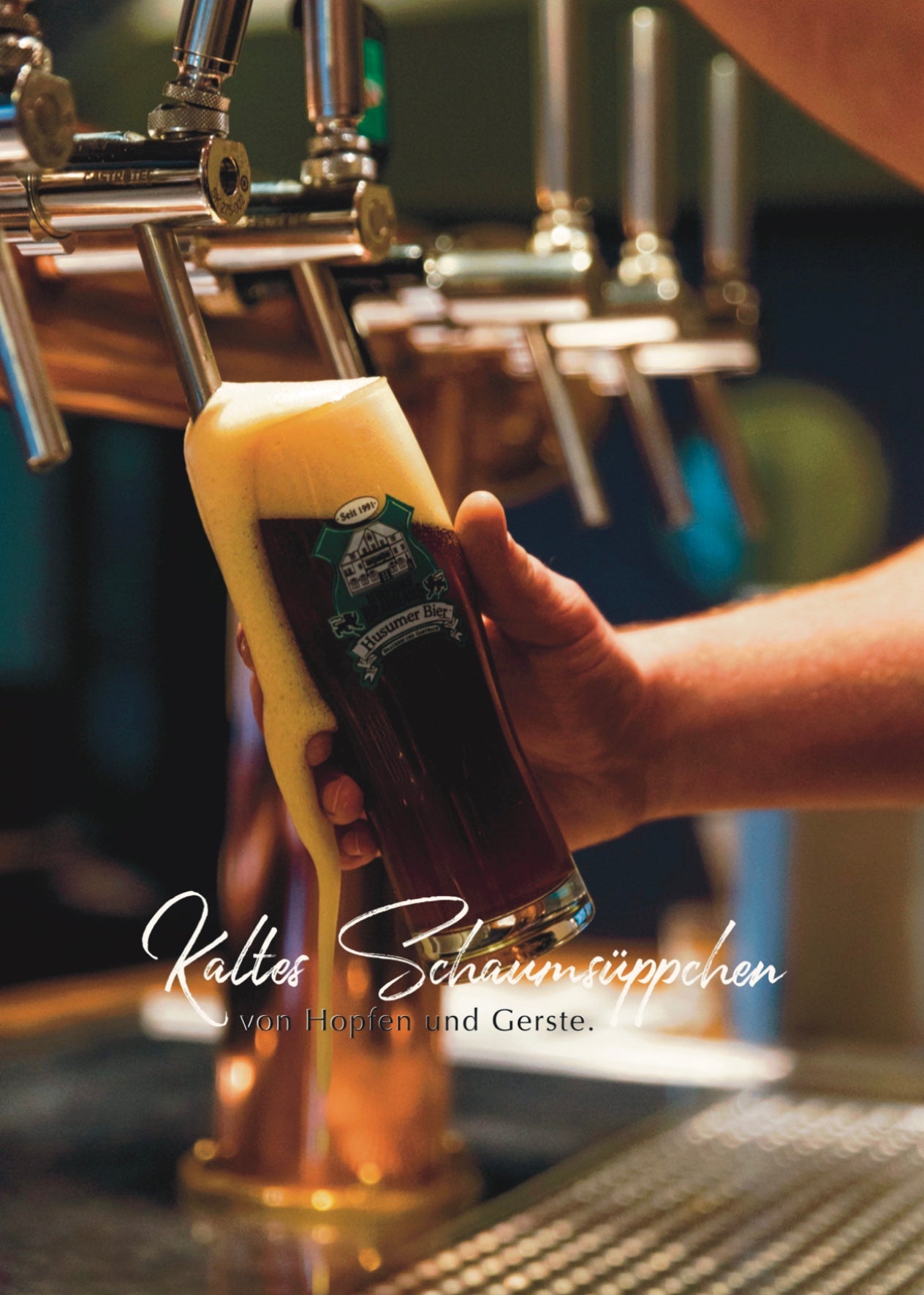 Das Foto zeigt den Zapfhahn aus dem Brauhaus. Man sieht das Glas in welches jemand das Bier zapft. Man erkennt nur die Hände die das Glas halten und den Zapfhahn bedienen. Auf dem Foto steht: Kaltes Schaumsüppchen von Hopfen und Gerste.
